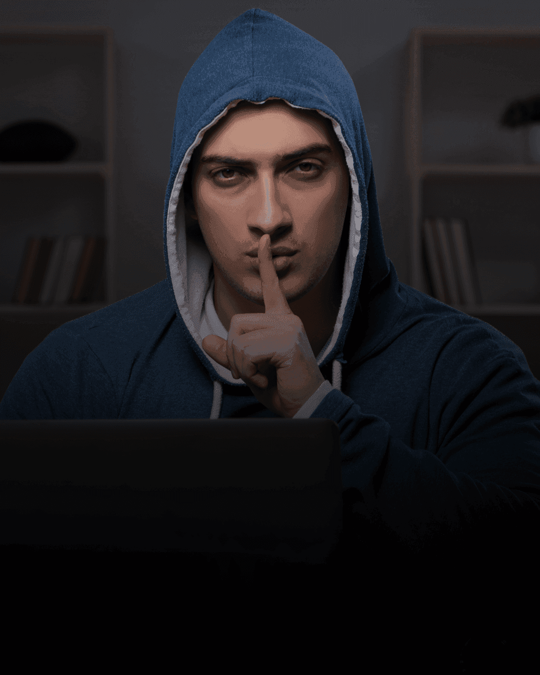 Computer hack in a hood in front of his laptop looking at the camera with his index finger on his lips